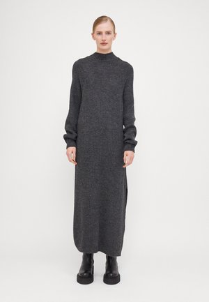 Long gray knit dress with a high neckline, ribbed details, and side slits. Worn with black platform boots, featuring long sleeves and textured fabric.