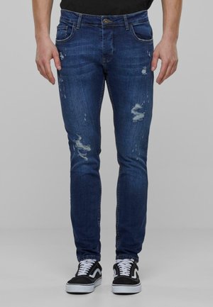 Vaqueros skiny de mezclilla azul con detalles desgastados, sutiles arrugas y diseño de cinco bolsillos. Combinados con zapatillas negras.