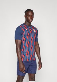 Kék és piros hullámmintás sportpóló rövid ujjakkal és kerek nyakkal, navy színű sorttal párosítva. Könnyű, lélegző anyagból készült.