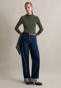 Olijfgroen aansluitend topje met een golfrand, gecombineerd met hoge donkere jeans. Bruin leren riem met een decoratieve sjaaldetail.