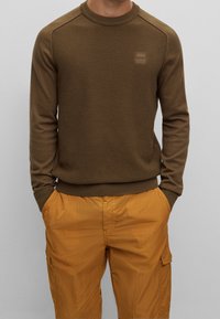 Brun texturerad stickad tröja med ribbade ärmslut och halsringning, med en fyrkantig logotyp-patch. Kombinerad med orange lätta cargoshorts.