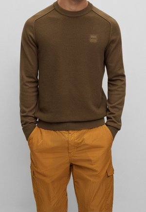 Brauner, strukturierter Strickpullover mit gerippten Säumen und Halsauschnitt, versehen mit einem quadratischen Logo-Patch. Kombiniert mit leichten orangenen Cargo-Shorts.