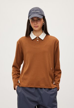 Jeune femme aux longs cheveux foncés portant une chemise marron à manches longues avec un col blanc, un pantalon bleu marine, des boucles d’oreilles créoles et une casquette bleue.