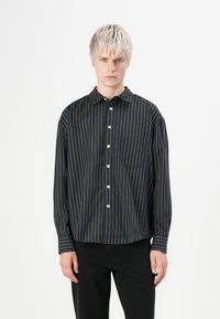 Young person with short blonde hair wearing a black long-sleeve button-up shirt with white vertical stripes and black pants.