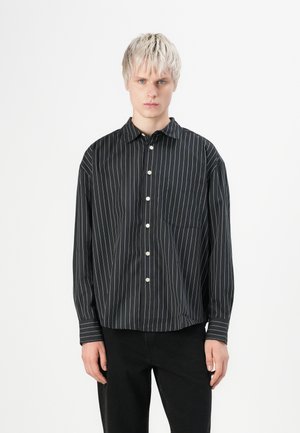 Jeune personne avec des cheveux blonds courts portant une chemise noire à manches longues avec des rayures verticales blanches et un pantalon noir.