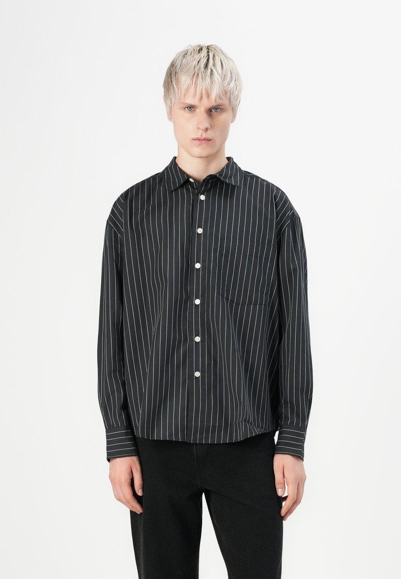 Young person with short blonde hair wearing a black long-sleeve button-up shirt with white vertical stripes and black pants.