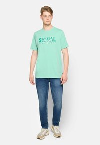 Camiseta de algodón verde claro con el texto gráfico "SIGNAL SINCE 1967" en verde más oscuro, combinada con jeans azules y zapatillas beige.