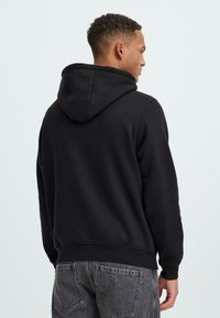 Juodas gaubtas megztinis, pagamintas iš minkštos medžiagos, su raišteliu reguliuojamu gaubtu, gipiūrinėmis rankovėmis ir liemeniu; derinamas su tamsiai pilkais džinsais.