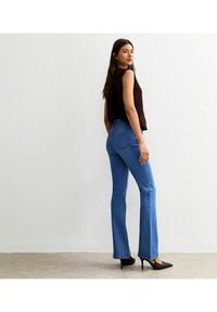 Donna con top senza maniche marrone scuro, jeans a vita alta svasati blu e scarpe con tacco nere, in piedi di lato contro un muro bianco.