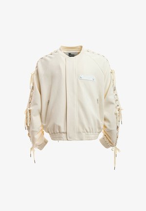 Joven con rastas cortas lleva una chaqueta bomber de color marfil, camisa de malla clara, pantalones cortos beige, calcetines blancos y zapatos beige texturizados.