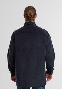 Mann mit braunen Haaren, die zu einem Dutt gebunden sind, trägt eine dunkelblaue Cordjacke und wird von hinten vor einem schlichten hellen Hintergrund gezeigt.