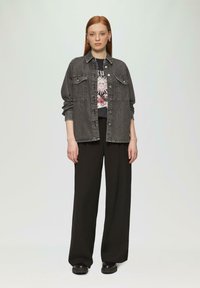 Zwarte denimjack met twee voorzakken over een graphic t-shirt met een zonafbeelding. Gekleed met brede zwarte broek en chunky zwarte schoenen.