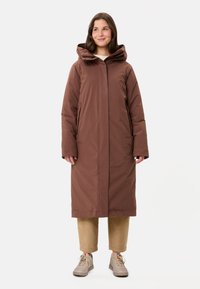 Manteau long marron avec capuche, doté d'une fermeture éclair sur le devant, de poches latérales et d'une texture lisse. Porté sur un pantalon en velours côtelé beige et des chaussures claires.