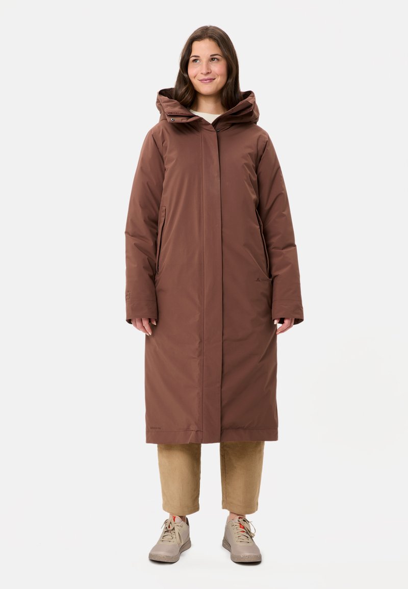 Manteau long marron avec capuche, doté d'une fermeture éclair sur le devant, de poches latérales et d'une texture lisse. Porté sur un pantalon en velours côtelé beige et des chaussures claires.