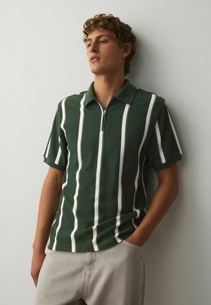 Young man with curly hair wearing a green polo shirt with white vertical stripes and light gray pants, standing against a plain white wall.