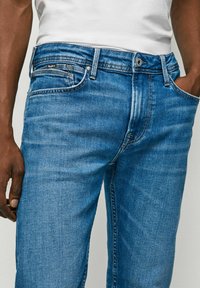 Jeans din denim albastru, cu o croială slim, având un design clasic cu cinci buzunare, închidere cu buton metalic și o ușoară decolorare pe material.
