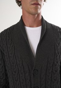 Donkergrijze gebreide cardigan met een sjawl-kraag, kabelbreipatroon en twee zwarte knopen. G gedragen over een witte T-shirt.