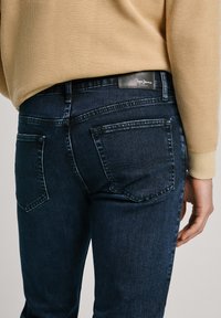 Temno modri jeans z ožjim krojem, ki imajo dve zadnji žepek in črn usnjen logotip na pasu. Zgoraj je pletena bež pulover.