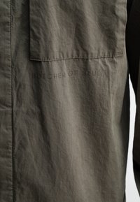 Korte mouwen, olijfgroene shirt gemaakt van een geweven stof. Heeft een borstzak en reliëftekst "BUTCHER OF BLUE" langs de zijnaad.