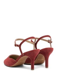 Červené semišové lodičky na vysokém podpatku s otevřenou zadní částí, páskem kolem kotníku a zaoblenou špičkou. Mají texturovaný povrch a štíhlý podpatek.