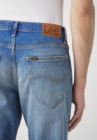 Modré džínové kalhoty s hnědým koženým štítkem s nápisem "Lee" na pasu, viditelným šitím a zadními kapsami s zakřivenými švy.