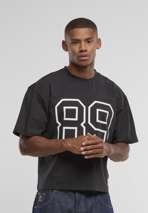 Young man with short hair wearing a black oversized t-shirt with white "89" print and blue denim jeans, standing against a plain background.