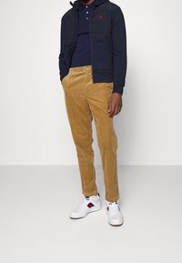 Sudadera con cremallera en color azul marino, camiseta polo de manga corta, pantalones de terciopelo marrón y zapatillas blancas con detalles en rojo y azul. Materiales suaves y ajuste a medida.