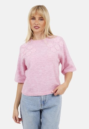 Vrouw met blond haar, gekleed in een roze korte mouwen trui met bloemenapplicaties en lichtblauwe hoge taille jeans, tegen een witte achtergrond.