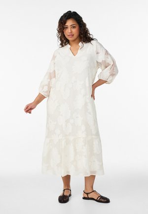 Kobieta w białej sukience midi w kwiaty z rękawami 3/4 zakończonymi bufkami oraz czarnych plecionych płaskich sandałach, stojąca na tle jednolitego białego tła.