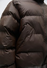 Brūns pūkains jakas ar matētu apdari, quilted dizainu un ar rāvējslēdzēju. Ietver sānu kabatu un stāvošu apkakli.