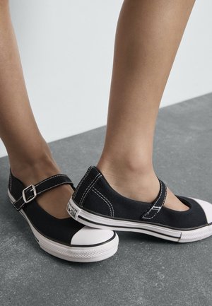 Czarne buty typu Mary Jane na płótnie z paskiem i białym gumowym noskiem. Charakteryzują się białym przeszyciem i gumową podeszwą.