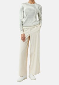 Camicia a maniche lunghe a righe in grigio chiaro e bianco, abbinata a pantaloni beige a gamba larga e sneakers bianche, in piedi con le mani in tasca.