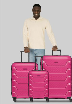 Hombre sonriente con suéter beige y jeans sostiene las asas de tres maletas rígidas de color rosa brillante, de diferentes tamaños y con ruedas.