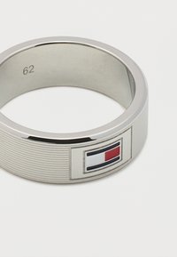 Ασημένιο μεταλλικό δαχτυλίδι με λεπτές οριζόντιες αυλακώσεις, με ορθογώνιο λογότυπο που διαθέτει κόκκινες, λευκές και μπλε ενότητες σε λευκό φόντο.