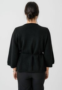 Cardigan negru, tricotat, cu talie legată, mâneci trei sferturi și o croială lejeră. Prezintă un decolteu rotund și o textură subtilă.