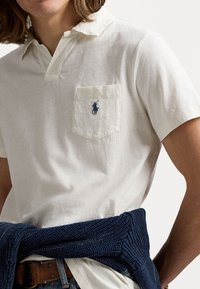 Polo shirt bianco a maniche corte con colletto aperto e taschino sul lato sinistro con un piccolo logo del cavallo ricamato in blu. Avvolto in un maglione blu navy.