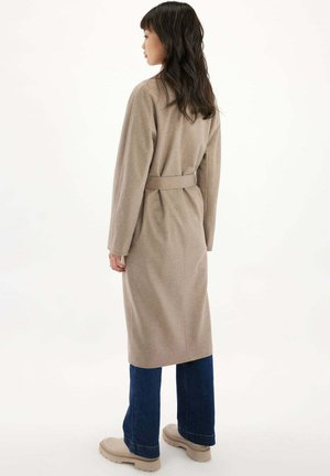 Frau, die nach links blickt und einen langen beigen Mantel mit Gürtel, dunkelblaue Jeans und beige Schuhe mit klobiger Sohle vor einem schlichten weißen Hintergrund trägt.