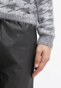 Mano appoggiata accanto a una gonna in pelle nera, indossando un maglione grigio in maglia pelosa con motivo testurizzato e polsini a costine.