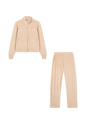 Chándal de terciopelo beige que cuenta con una chaqueta con cremallera, cuello alto y bolsillos laterales, combinado con pantalones ajustados y rayas blancas.