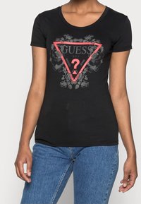 Svart bomulls T-shirt med rund halsringning och korta ärmar. Har en röd triangulär logotyp med blommönster och texten "GUESS" och "WASHED JEANS."