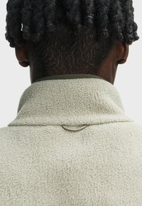 Widok z tyłu osoby z krótkimi, skręconymi czarnymi włosami ubranej w szarą, teksturowaną bluzę polarową z kołnierzem obszytym na zielono i małą pętelką.