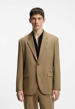 Jeune homme aux cheveux courts et foncés portant une veste de costume beige par-dessus un haut noir à fermeture éclair, sur un fond blanc uni.
