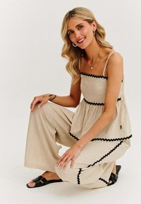 Světle béžový overal s černým vlnitým lemem a širokými nohavicemi. Vyroben z texturované tkaniny, s tenkými ramínky a přiléhavým korzetem.