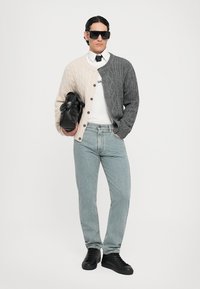Tvåfärgad kabelstickad cardigan i beige och grått, kombinerad med en vit skjorta, ljusblå jeans och svarta sneakers. Modellen håller en svart väska.