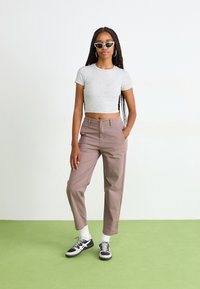 Grijze cropped t-shirt, lichtbruine hoge taillebroek en zwarte sneakers met witte accenten. Model draagt kattenoog zonnebril en creolen.