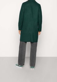 Grüner Mantel mit geradem Schnitt, langen Ärmeln und Kragen, kombiniert mit grauen Hosen mit weitem Bein und grünen Turnschuhen.