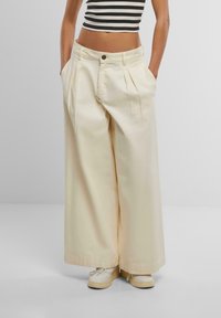 Wide-leg beige cotton trousers with pleats and side pockets, featuring a button closure, paired with white sneakers.