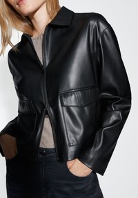 Giubbotto corto in pelle nera con colletto, chiusura con zip e due tasche frontali. Tessuto liscio e design aderente.