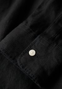 Puño de camisa de lino negro con un botón blanco. Presenta costuras visibles y una textura suave. El botón tiene un patrón floral.