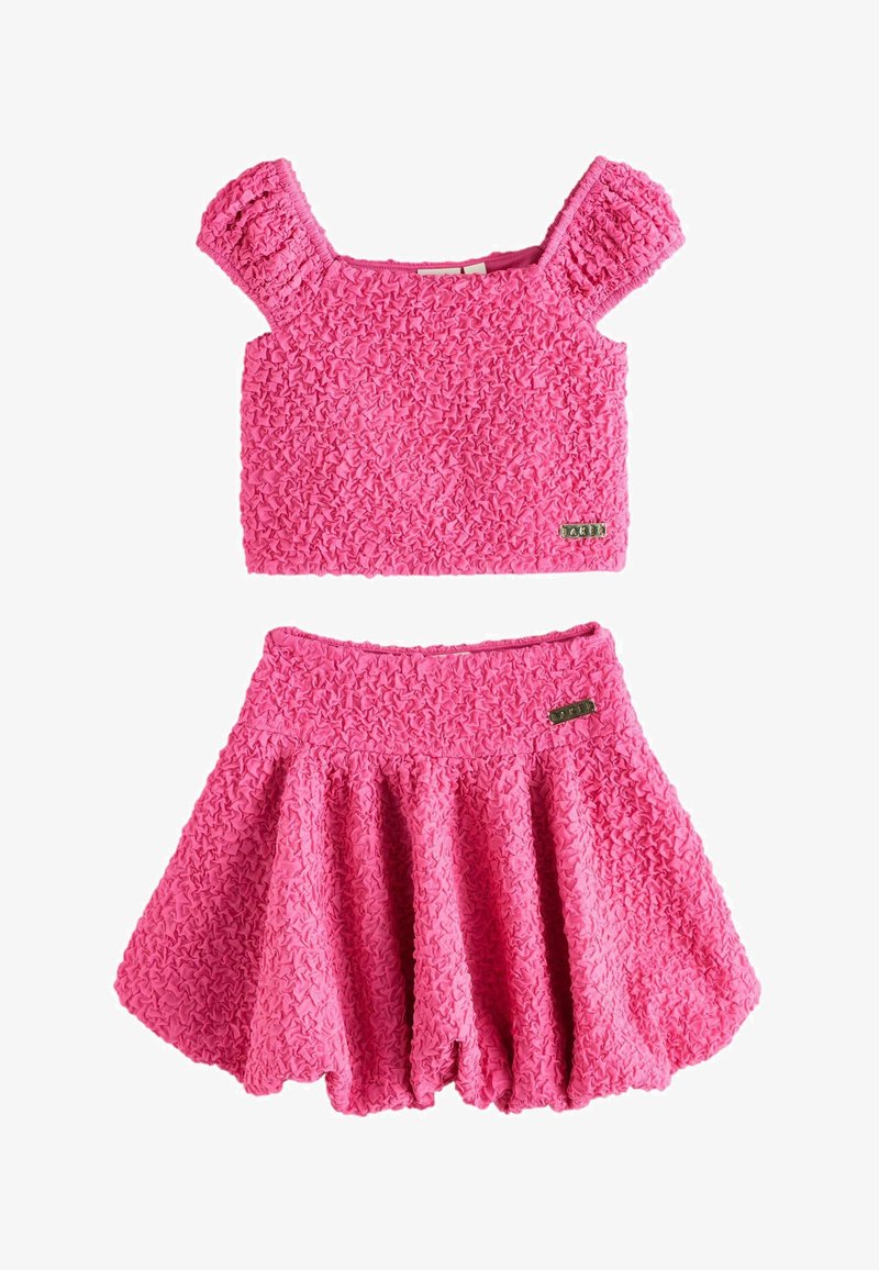 Rosa strukturiertes zweiteiliges Outfit mit kurz puffigen Ärmeln am Croptop und ausgestelltem Rock, beide mit kleinem metallischem Markenzeichen versehen.